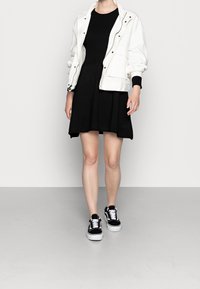Veste bomber blanche sur une robe noire à côtes, associée à des baskets noires et blanches. Tissu lisse avec des accents contrastants et une silhouette ajustée.