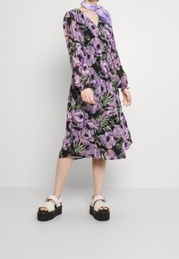 Květinové šaty s dlouhými rukávy, s černým pozadím a fialovými a zelenými květinami, s přiléhavým pasem; spárovány s bílými masivními sandály.