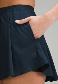 Falda atlética azul marino con diseño acampanado, fabricada en un tejido ligero, con cintura elástica y un bolsillo lateral. Presenta un logo en el dobladillo inferior.