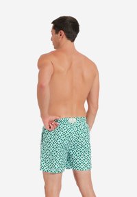 Costumi da bagno verdi con un motivo geometrico bianco, vita elastica e una tasca posteriore con etichetta; il tessuto appare leggero e dalla texture particolare.