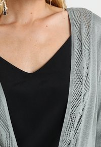 Světlé šedé pletené kardigan s geometrickým vzorem na okrajích, vrstvený přes černý top s výstřihem do V. Tkanina vypadá měkce a lehce.