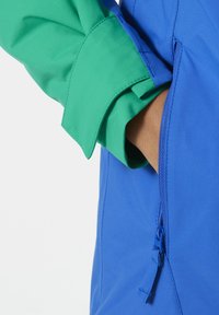 Wielokolorowa kurtka z niebieską podstawą i zielonymi mankietami. Posiada zasuwaną kieszeń z boku oraz gładki, lekki materiał.