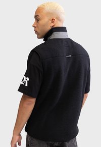 Gilet en polaire noir avec un col gris, présentant un col montant, un tissu texturé et un petit détail en boucle à l'arrière. Manches courtes en dessous.