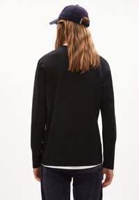 Schwarzes Langarmshirt mit rundem Ausschnitt, unter einem weißen Shirt getragen, kombiniert mit blauen Hosen und einer blauen Mütze. Glatte Textur.