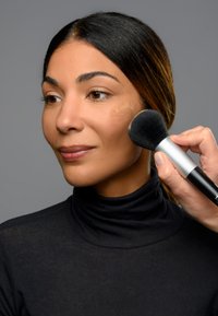 Die Anwendung von Make-up wird fortgesetzt, während mit einem schwarzen Make-up-Pinsel die Wange einer Frau betont wird. Ihr Gesicht ist mit einer hellen Foundation versehen und der Hintergrund ist grau.