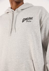Light gray hoodie made of soft material, featuring a front pocket. Black "Groove in Bloom" logo printed on the chest. Adjustable drawstring hood.