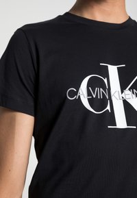 Svart bomullströja med en stor vit "CK"-logotyp och hela "CALVIN KLEIN"-texten. Standard rund hals och korta ärmar.