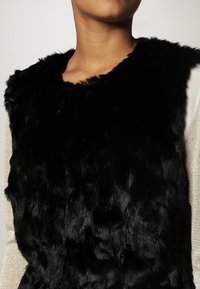 Musta tekoturkisliivi, jossa on korkea kaulus, pehmeä, teksturoitu pinta ja tyylikäs muotoilu, jota vasten on vaaleampi vaatekappale alla.