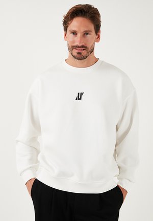 Biała bluza z małym czarnym logo na piersi, okrągłym dekoltem i luźnym krojem, wykonana z miękkiego materiału.