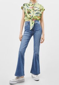 Kortärmad, knuten blus med blommönster i ljusgrönt, ihop med högmidjade blå utsvängda jeans och vita kanvas sneakers.