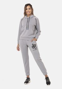 Grauer Hoodie und Jogginganzug aus weichem Stoff, mit einer Fronttasche, Kapuze mit Kordelzug und Logodetails auf der Hose. Schwarze Sneakers runden das Outfit ab.