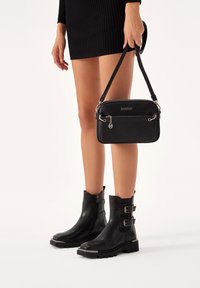 Black leather handbag with zip, silver hardware, and a front pocket. Worn with black ankle boots featuring buckles and a chunky sole.