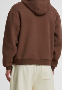 Schokoladenbraun Kapuzenpullover in lässigem Schnitt, mit Kapuze, gerippten Bündchen und Saum, aus weichem Material. Getragen mit hellen Hosen.