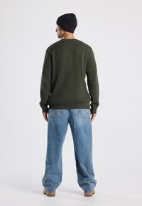 Hombre con gorro negro, suéter de canalé verde oscuro, jeans azules holgados y botas color dátil, de pie de espaldas sobre un fondo blanco.