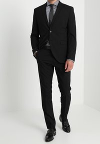 Czarny, dopasowany garnitur o wąskim kroju, z jednorzędową marynarką na jeden guzik, klapami z wycięciem i dopasowanymi spodniami. Połączony z szarą koszulą i krawatem.