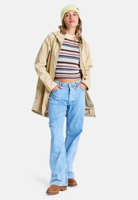 Beige wasserdichte Jacke, gestreifter cropped Pullover, hellblaue Schlagjeans und braune Stiefeletten. Gelbe Strickmütze auf dem Kopf.