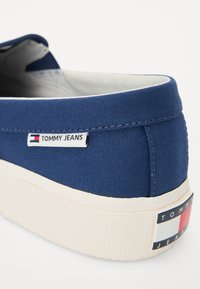 Zapatilla de lona en color navy con suela de goma blanca, un diseño cosido y una etiqueta de marca con acentos en rojo, blanco y azul en el talón.