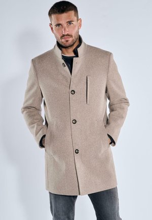 Beiger Wollmischmantel mit hohem Kragen, Frontknöpfen, einer Brusttasche und Seitentaschen, kombiniert mit dunklen Jeans. Glatte Textur.