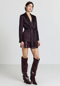Blazer borgogna con cintura a fascia, abbinato a una gonna mini in pelle coordinata e stivali al ginocchio, caratterizzati da una texture lucida e punte affusolate.