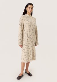 Beiges, geripptes Kleid mit hohem Kragen, langen Ärmeln und schwarzen Sprenkeln. Kombiniert mit dunkelbraunen Ballerinas.