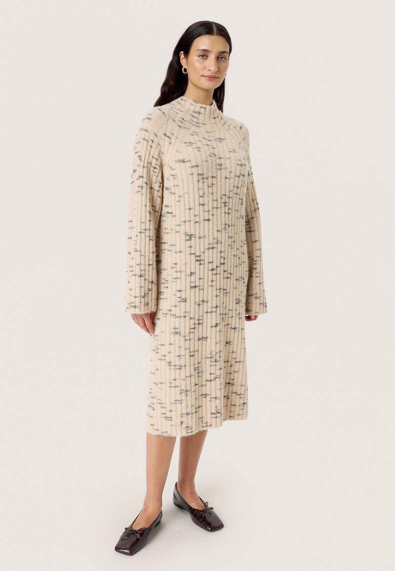 Beiges, geripptes Kleid mit hohem Kragen, langen Ärmeln und schwarzen Sprenkeln. Kombiniert mit dunkelbraunen Ballerinas.