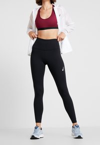 Bordowy stanik sportowy, lekka biała kurtka, czarne legginsy z wysokim stanem oraz jasnoniebieskie buty do biegania z detalem logo.