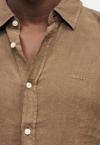 Κοντινό πλάνο ενός ατόμου που φοράει ένα καφέ πουκάμισο με ανοιχτά κουμπιά, με γιακά και κεντημένο λογότυπο "BOSS" στο στήθος.