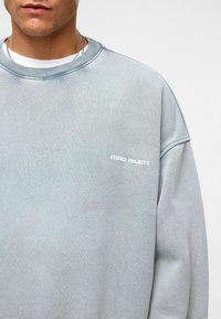 Lichtblauwe sweatshirt gemaakt van zacht materiaal. Kenmerkt zich door een geribde ronde hals en een wit "ESSENCE PROJECT" logo op de borst.