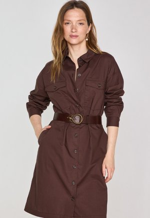 Bruine shirtjurk met lange mouwen, knopen aan de voorkant en twee borstzakken, in de taille cinched met een donkere riem met een decoratieve gesp.