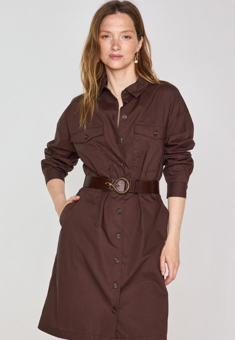 Abito camicia marrone con maniche lunghe, chiusura a bottoni frontale e due tasche sul petto, cinched in vita con una cintura scura con fibbia decorativa.