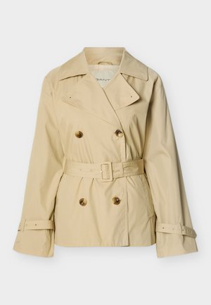Trench-coat beige à double boutonnage avec larges revers, taille ceinturée, poignets boutonnés et poches plaquées sur fond blanc.