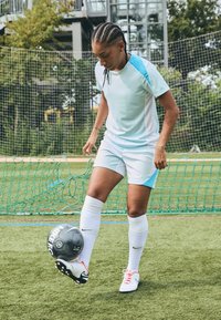 Hele sinine spordipoliis valgete paneelidega, koos valgete lühike pükstega. Mängija tasakaalustab musta Nike jalgpalli ühel jalal, kandes valgeid sokke ja kummikuid.