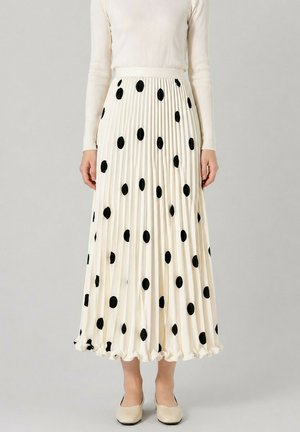 Femme portant une jupe midi plissée blanche à gros pois noirs, associée à un haut blanc à manches longues et des chaussures beiges.