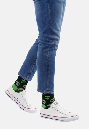 Person iført blå jeans, sorte sokker med grønne alienansigter og hvide lave Converse-sneakers.