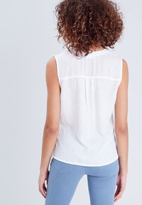 White sleeveless blouse with subtle floral patterns and a smooth texture. Features a gently curved hem and a central back pleat.