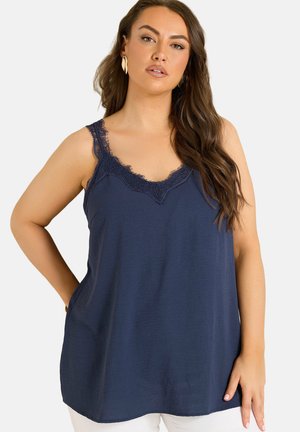 Vrouw met lang bruin haar draagt een marineblauwe mouwloze top met kanten rand en witte broek, poseert met één hand op haar heup.