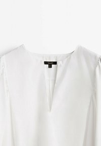 Blouse blanche à col en V, manches courtes et détails plissés subtils. Fabriquée en tissu lisse, avec une étiquette au centre du dos.
