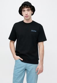 Μαύρο βαμβακερό μπλουζάκι με κοντά μανίκια, με μπλε κεντημένο λογότυπο "Dickies" στο στήθος, συνδυασμένο με ανοιχτόχρωμο τζιν και μαύρο καπέλο τύπου bucket.