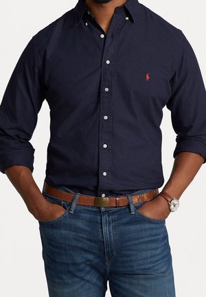 Mężczyzna w granatowej koszuli na guziki z czerwonym logo, niebieskich dżinsach, brązowym skórzanym pasku i zegarku na rękę, z rękami włożonymi do kieszeni.