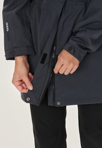 Mörkgrå vattentät jacka med dragkedja och tryckknappar, med en texturerad tyg och justerbara ärmar. Närbild av hand på dragkedjan.