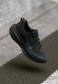 Μαύρο αθλητικό παπούτσι με υφητή επάνω επιφάνεια, διαθέτοντας μια λεία, στρογγυλή σόλα και κορδόνια, με λεπτομέρεια ενός διακριτικού γκρι λογότυπου.