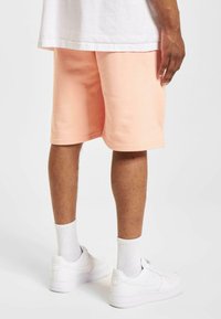 Shorts en coton pêche avec une coupe décontractée, texture lisse et longueur au genou ; associés à des baskets blanches et des chaussettes blanches côtelées.