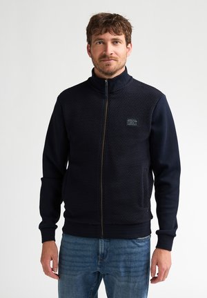 Mann mit kurzen braunen Haaren und Bart, trägt eine strukturierte schwarze Reißverschlussjacke und blaue Jeans, steht vor einem schlichten weißen Hintergrund.