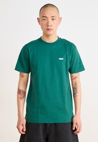 Tricou verde din bumbac cu mâneci scurte, având un logo alb "OBEY" pe pieptul stâng. Textură netedă, croială lejeră.