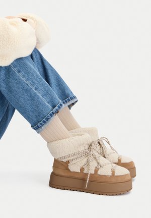 Masywne brązowe botki na platformie z grubą kremową podszewką z polaru i sznurowadłami, noszone z beżowymi prążkowanymi skarpetami i podwiniętymi niebieskimi dżinsami na białym tle.