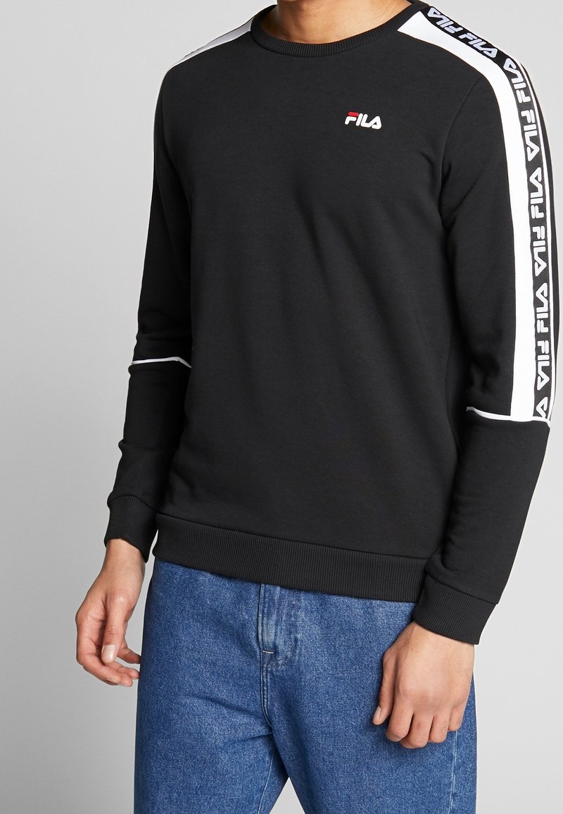 Homme portant un sweat-shirt FILA noir avec des bandes blanches à logo sur les manches et un jean bleu, debout devant un fond gris.
