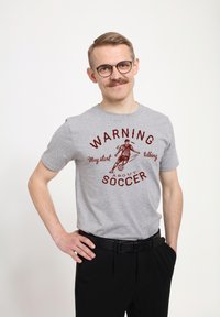 Camiseta gris con texto y gráficos rojos que dice "ADVERTENCIA Puede empezar a hablar sobre FÚTBOL". Mangas cortas, cuello redondo, corte casual.