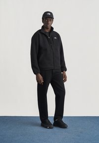 Άνδρας που στέκεται σε μπλε χαλί, φορώντας ολοκληρωμένη μαύρη εμφάνιση, συμπεριλαμβανομένου fleece μπουφάν, καπέλου, παντελονιού και αθλητικών παπουτσιών με λευκές λεπτομέρειες λογότυπου.