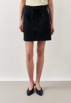 Žena nosiaca čiernu sukňu po kolená s predným viazaním a čierne špicaté topánky, stojaca pred jednoduchým svetlým pozadím.