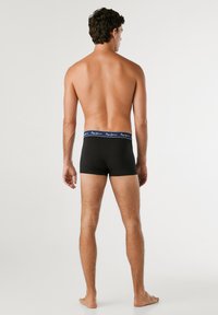 Schwarze Baumwoll-Boxershorts mit einem blau-weißen Markenbund, ausgestattet mit einem passgenauen Design und mittellanger Beinlänge. Von hinten betrachtet.